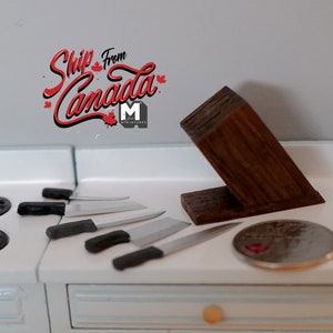 Puede incluir: Un bloque de cuchillos de madera en miniatura con cinco cuchillos en miniatura sobre una encimera blanca. El bloque de cuchillos es marrón y tiene una veta de madera natural. Los cuchillos son plateados con mangos negros. Hay una pequeña tabla de cortar redonda de plata a la derecha del bloque de cuchillos.