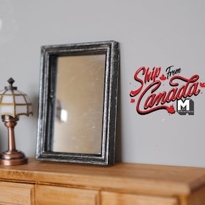 May include: A black framed mirror with a silver finish sits on a wooden dresser with a lamp with a white and gold shade. The wall behind the dresser has the text "Ship from Canada" in red lettering with maple leaf accents.