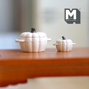Può includere: Due pentole in ceramica bianca a forma di zucca con coperchi. La pentola più grande ha due manici e la pentola più piccola ha un manico. Entrambe le pentole hanno un pomello marrone sul coperchio.