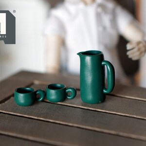 Op de afbeelding: Een set miniatuur groen keramiek keukengerei, inclusief een kan en twee kopjes. De set staat op een houten tafel.