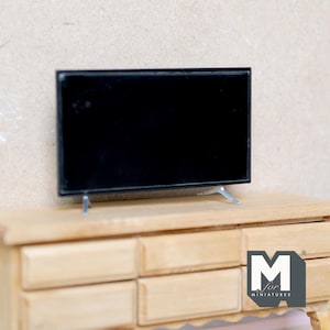 May include: A miniature black flat-screen TV with silver legs rests on a light wood dresser. The TV has a black screen and a thin black frame. The dresser has six drawers and a natural wood finish. The image is of a miniature item.