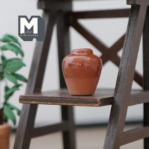 May include: A small, terracotta-colored ceramic pot sits on a wooden shelf. The pot is round with a slightly flared lip and a smooth, glossy finish.