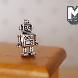 May include: A small, silver robot figurine with a boxy head, round eyes, and three circular details on its chest. The robot stands on a wooden surface. The logo "M for miniatures" is in the background.