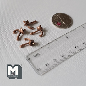 May include: Four small, copper-colored miniature hooks with a coin and a ruler for scale. The hooks have a curved design with a circular top and a small peg. The ruler shows measurements in inches.