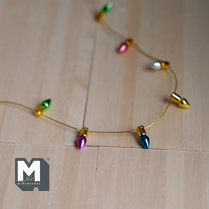 May include: A string of miniature Christmas lights with green, pink, blue, and silver bulbs. The lights are strung on a gold wire and are hanging on a wooden surface.