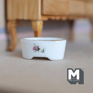 以下が含まれることがあります： 金色の縁取りとピンクのバラの模様が施された、小さな白い磁器製の皿。皿は楕円形で、3つの小さな足がついています。