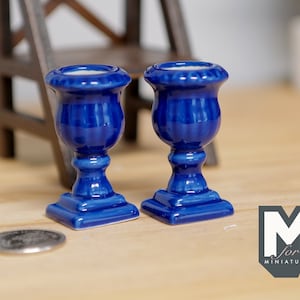 May include: Two cobalt blue miniature urns with a glossy finish. Each urn has a square base, a pedestal, and a flared top. The urns are displayed on a light-colored surface. A coin is in the foreground.