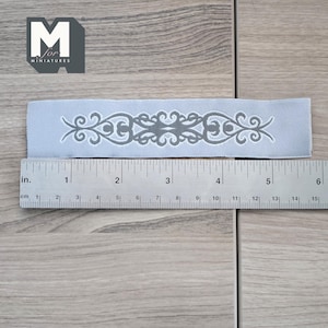 May include: A gray fabric trim with a decorative silver design. The trim is approximately 5 inches long.