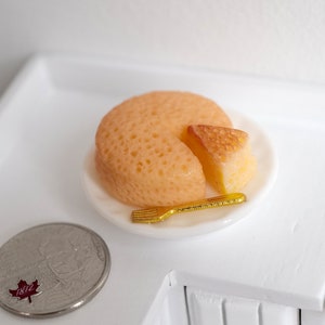 May include: A miniature yellow cake with a slice cut out, served on a white plate with a gold plastic fork. A Canadian quarter coin is in the foreground.