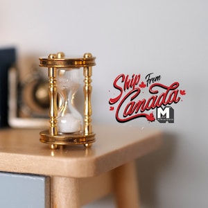 May include: A gold hourglass with white sand sits on a wooden table. The hourglass is about 3 inches tall. The background is a white wall with the text "Ship from Canada" in red.