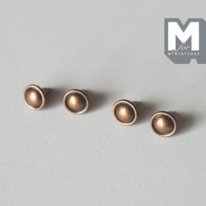 May include: Four small, round, copper-colored metal studs with a raised center dome and a flat rim. The logo M for Miniatures is visible in the upper right corner. These circular fasteners feature a metallic finish and a pointed back attachment.
