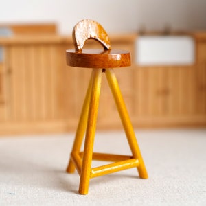 Op de afbeelding: Een houten kruk met een ronde zitting en een gebogen rugleuning. De kruk is gemaakt van lichtbruin hout en heeft een gele afwerking.