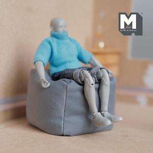May include: A grey fabric beanbag chair with a person sitting on it. The person is wearing a light blue sweater and blue jeans. The beanbag chair is a miniature piece of furniture.