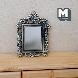 May include: A miniature ornate gold-toned mirror with a black frame and a reflective surface. The mirror is on a wooden surface.
