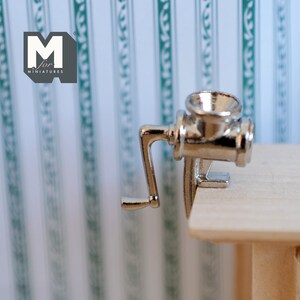May include: A silver miniature hand-crank meat grinder with a handle on the side. The grinder is sitting on a wooden surface.