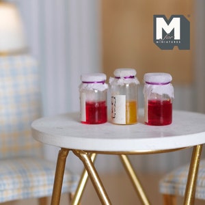 Peut inclure: Trois petits pots en verre avec des couvercles blancs et des finitions violettes. Les pots sont remplis de liquides rouges et jaunes. Les pots sont posés sur une table blanche avec des pieds dorés. Les pots sont étiquetés "M for Miniatures".