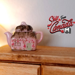 May include: A miniature pink teapot shaped like a cottage with a brown thatched roof and red berry accents. The teapot is sitting on a wooden surface.