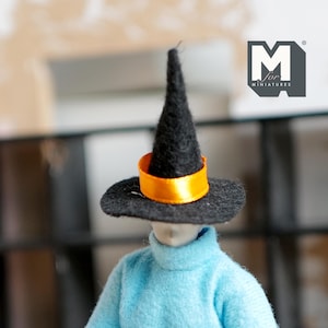 Op de afbeelding: Een miniatuur zwarte heksenhoed met een feloranje band, gedragen door een pop met een lichtblauwe coltrui. De hoed heeft een brede rand en een hoge, puntige kroon. De afbeelding bevat de tekst "M for MINIATURES".