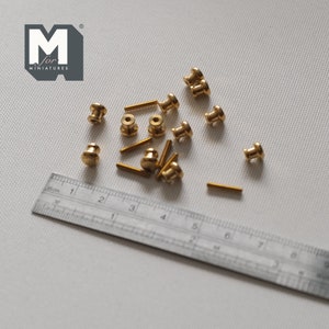 May include: A close-up of a group of 12 gold-toned metal cabinet knobs, some with screws, scattered on a white surface. A ruler is visible in the bottom left corner of the image.