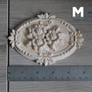 May include: An oval-shaped light-colored wooden carving featuring three central flowers surrounded by leaves and decorative border elements. A metal ruler placed below the carving shows it measures approximately 4.5 inches in width.
