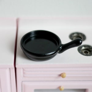 May include: A black miniature frying pan with a handle sits on a pink toy stovetop.