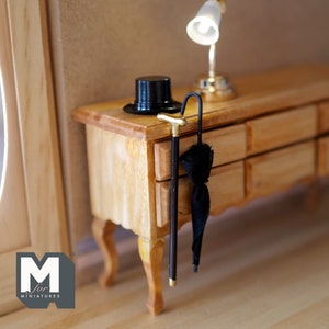 Op de afbeelding: Een miniatuur houten dressoir met drie laden, een zwarte hoge hoed, een zwarte paraplu en een wandelstok met een gouden punt. Op het dressoir staat een lamp met een witte kap.