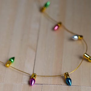 May include: A delicate gold-toned wire necklace featuring small, multi-coloured metallic light bulb charms in shades of green, pink, silver, blue, and yellow. Each charm is attached to the thin, flexible gold-coloured wire chain.