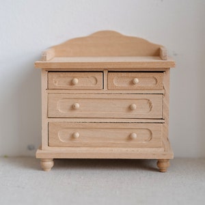 Op de afbeelding: Een kleine, onafgewerkte houten commode met vier lades en een decoratieve bovenkant. De commode heeft afgeronde knoppen en staat op vier kleine, gedraaide poten. De houtnerf is zichtbaar. Ideaal voor poppenhuizen of knutselprojecten.