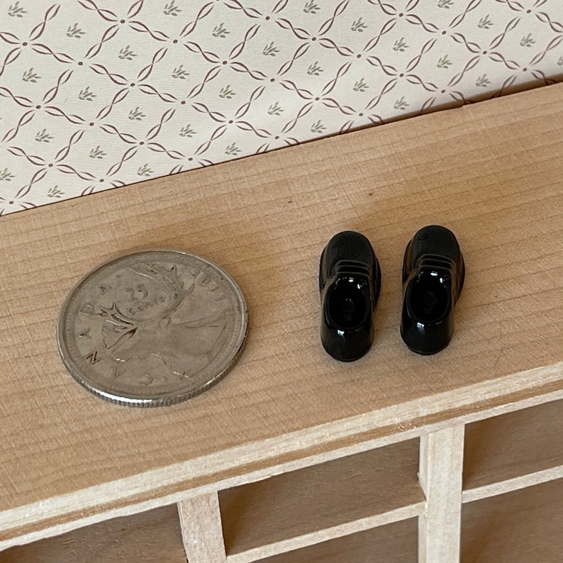 Pu&ograve; includere: Un paio di scarpe nere in miniatura per bambole. Le scarpe sono grandi circa come una moneta canadese da 25 centesimi.