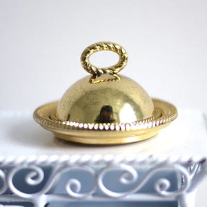 May include: A gold-toned serving dish with a lid and a decorative handle. The dish is on a white surface with a decorative design.