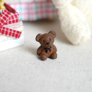 May include: A small brown teddy bear with a black bow tie. The bear is sitting on a white fabric surface.