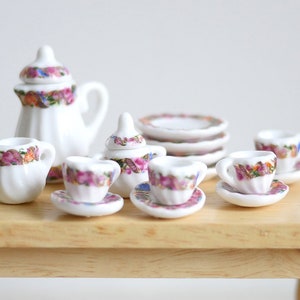 Könnte beinhalten: Ein Miniatur-Teeservice aus weißem Porzellan mit floralen Akzenten. Das Set beinhaltet eine Teekanne, einen Sahnekännchen, eine Zuckerdose, Tassen, Untertassen und Teller. Das Blumendesign zeigt rosa, lila und gelbe Blumen.