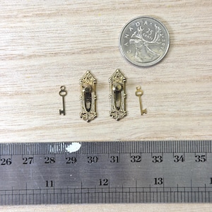 Peut inclure: Deux plaques de serrure miniatures en métal doré avec des volutes ornementales et deux petites clés assorties. Une pièce de 25 cents canadienne et une règle en métal sont incluses pour l'échelle, montrant une longueur d'environ 2,5 cm.