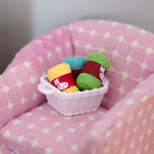 Op de afbeelding: Een miniatuur roze fauteuil met witte stippen houdt een klein wit mandje vast gevuld met kleurrijke garenbollen. De garenkleuren zijn onder andere geel, groen en blauw. Het mandje heeft een handvat en een decoratief ontwerp.