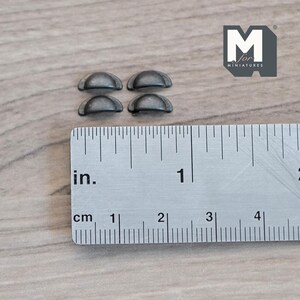 May include: Four small, antique-style metal drawer pulls, arranged in a 2x2 pattern, sit on a wooden surface next to a ruler. The ruler shows measurements in inches and centimeters. The drawer pulls are approximately 0.5 inches in diameter.