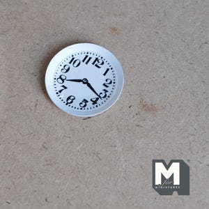 May include: A white clock face with black numbers and hands. The clock face is 2 inches in diameter and is made of paper.