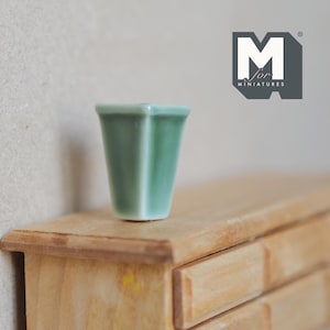 Op de afbeelding: Een kleine, groene keramische pot met een vierkante basis en een licht uitlopende bovenkant. De pot staat op een houten oppervlak.