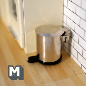 Puede incluir: Un cubo de basura en miniatura de plata con tapa y base negra. El cubo de basura está sobre una superficie de madera.