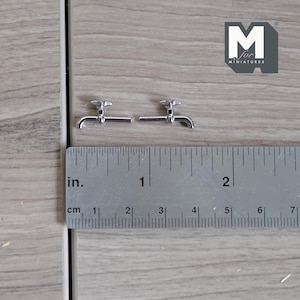 May include: Two silver miniature taps with a ruler for scale. The ruler measures 5 cm.