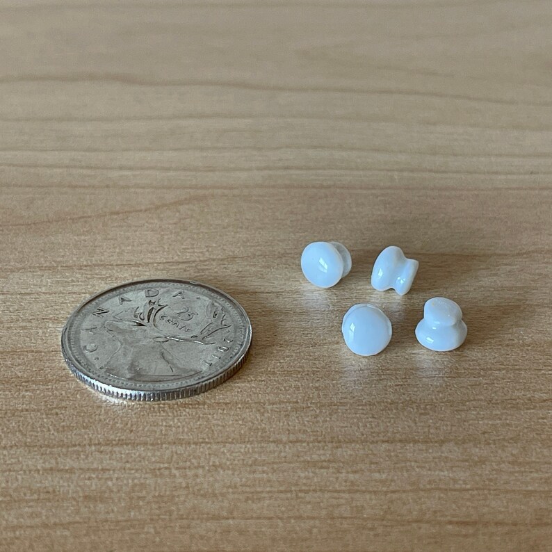 Może przedstawiać: Cztery białe ceramiczne gałki z gładką, zaokrągloną powierzchnią. Gałki są ułożone w p&oacute;łkole na jasnobrązowej drewnianej powierzchni.