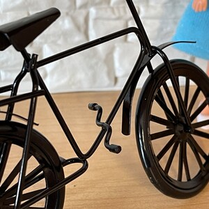 May include: A black metal bicycle model with a curved handlebar, a black seat, and two black wheels. The bicycle is on a wooden surface.