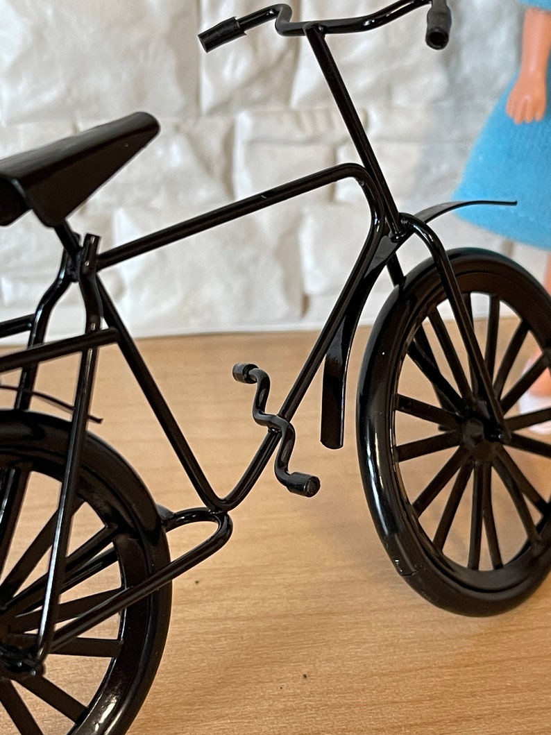 May include: A black metal bicycle model with a curved handlebar, a black seat, and two black wheels. The bicycle is on a wooden surface.