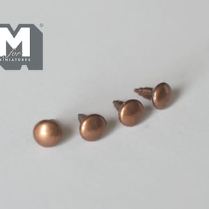 May include: Four small, round-headed upholstery nails with a metallic copper finish and threaded shanks. The nails are arranged in a row. A grey logo with the text M for MINIATURES is visible in the upper left corner of the image.