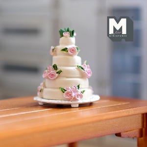 May include: A white wedding cake with pink and green frosting decorations. The cake is on a white cake stand. M for Miniatures is in the background.