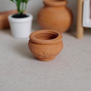 May include: A small, unglazed terracotta pot with a rounded body and a slightly flared rim. The pot has a textured surface with a simple embossed design. Other terracotta pots and a white potted plant are in the background.