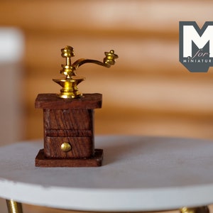 Könnte beinhalten: Eine Miniatur-Holzkaffeemühle mit einer goldenen Metallkurbel und einer Schublade. Die Mühle steht auf einem runden weißen Tisch.