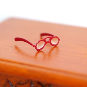 Op de afbeelding: Een kleine bril met een rood montuur en ovale glazen. De bril ligt op een houten oppervlak. Het montuur is felrood en de glazen zijn helder.