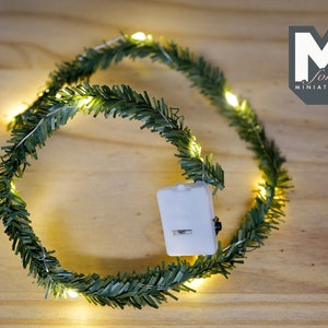 May include: A green garland with warm white LED lights wrapped around it. The garland is battery operated and has a small white battery pack attached to it. The garland is perfect for decorating a small space or adding a touch of festive cheer to a room.