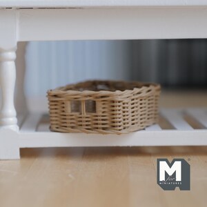 Op de afbeelding: Een kleine, geweven rieten mand met een rechthoekige vorm en een handvatachtige opening. De mand is lichtbruin en staat op een witte plank. De plank maakt deel uit van een witte tafel of kast. De mand is ontworpen voor miniatuurartikelen.
