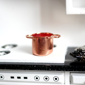May include: A miniature copper pot with two handles sits on a white countertop with a black hob in the background.